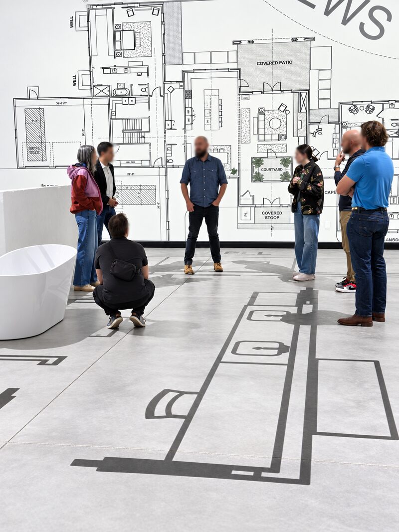 A group standing on a full-scale projected floor plan in front of a wall-sized floor plan drawing